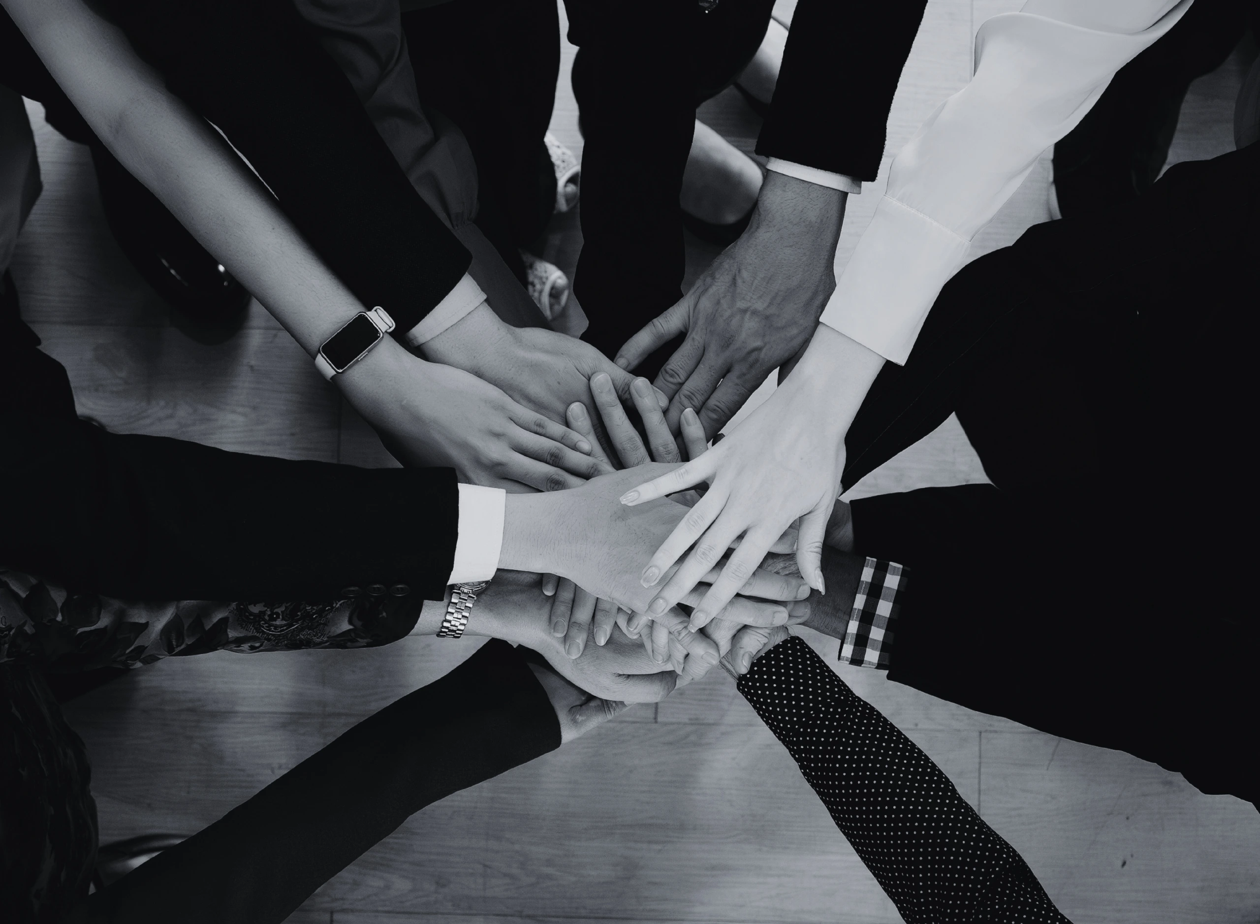 A high-angle, black and white photograph of diverse hands layered together in a central stack, symbolizing teamwork and unity.