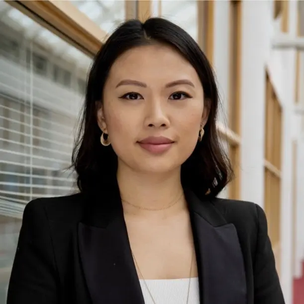 Rachel Jao smiling in a professional headshot wearing a black blazer and hoop earrings with an office window background.